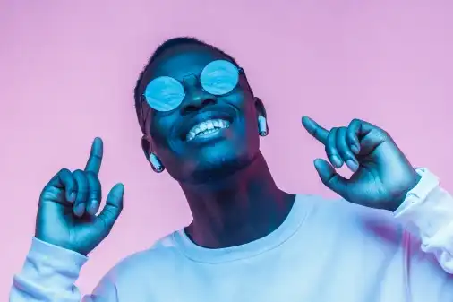 Happy man wearing hip rounded glasses and putting both fingers in the air pointing at his earbuds