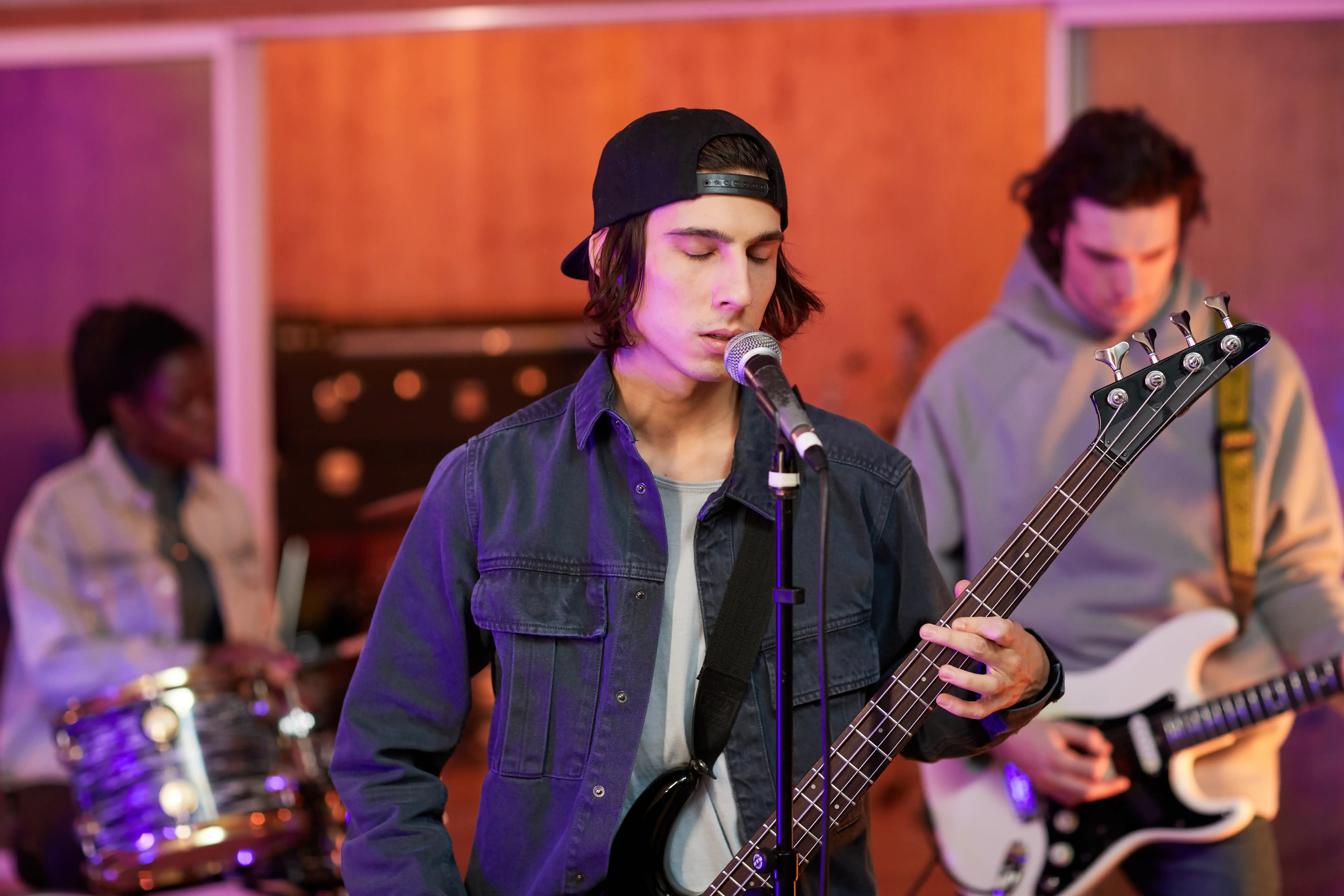 Medium shot of a singer in a rehearsal room playing a bass guitar, with in the background a guitarist and a female drummer.